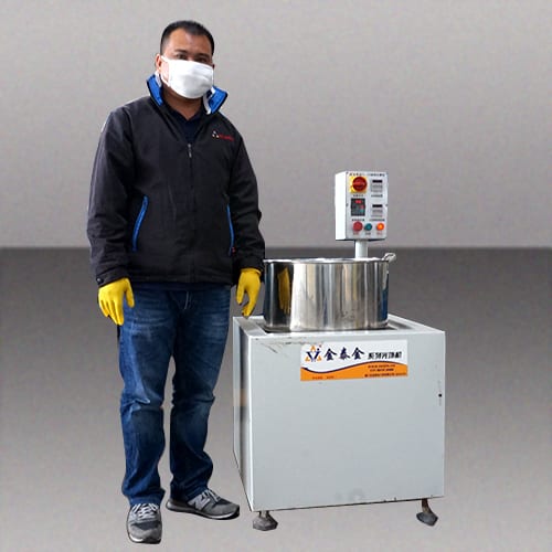 The image shows a man wearing a face mask and gloves standing next to a stainless steel mixing machine. He is wearing a black jacket, blue jeans, and black shoes. The background of the image is a wall