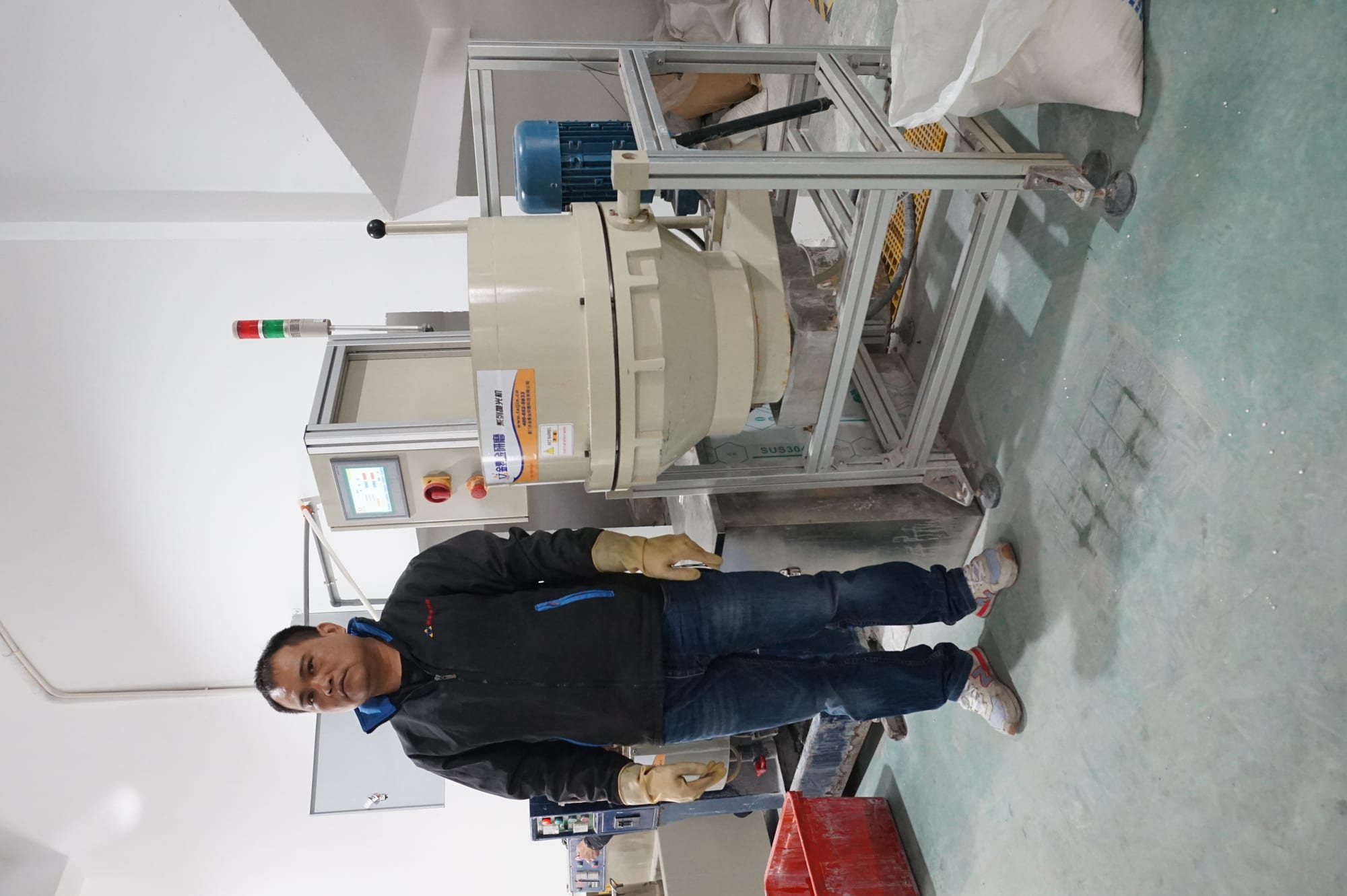 The image shows a man standing in front of a machine in a room. He is wearing a black jacket and blue pants, and is holding an object in his hand. On the right side of the image, there is a red box on